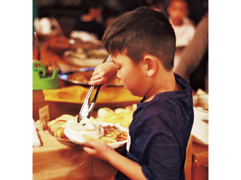 『こども食堂』で、テーブルに並べられた大皿料理から好きな食べ物を自分で皿によそう男の子。リンゴ、好きなんだね!