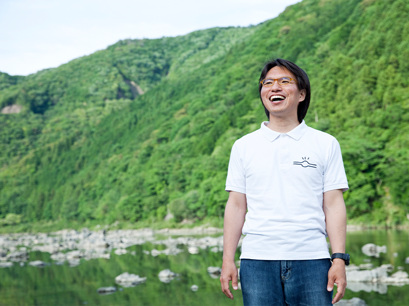 四万十川を背景に、笑顔の佐々倉さん。『いなかパイプ』のロゴ入り、白ポロシャツがよくお似合い!