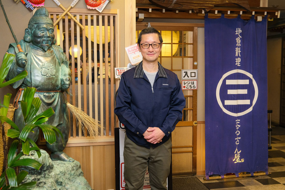 まるさん屋の社長が笑顔で立っている様子