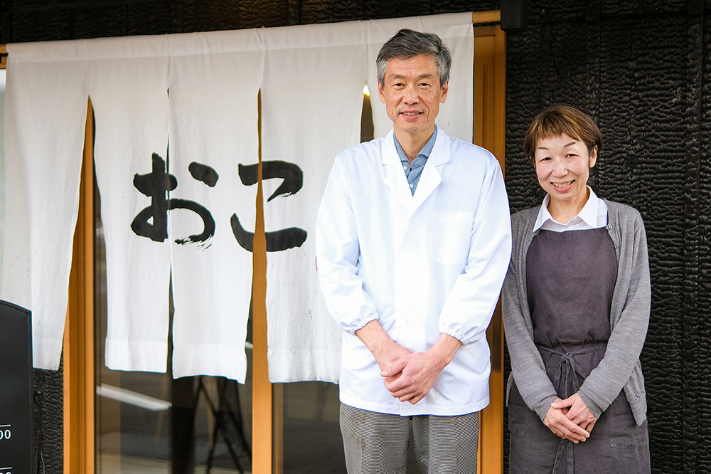 暖簾前に並ぶ、「美食ダイニング おこ」店主 島田さんご夫婦