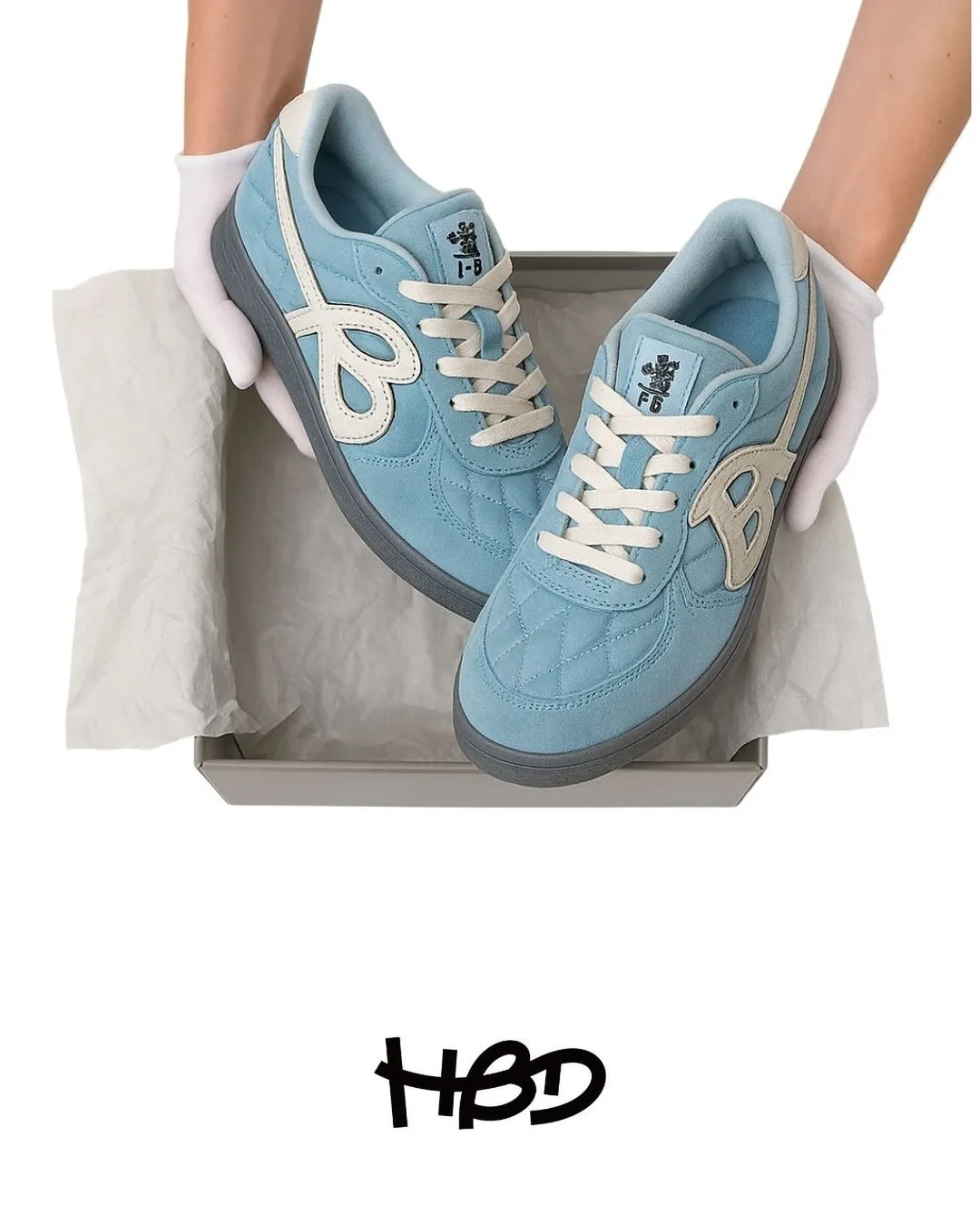 水色のスニーカーが箱の中に置かれ、白い手袋をはめた手で持たれている写真です。スニーカーには、ブランドロゴや文字がデザインされています。