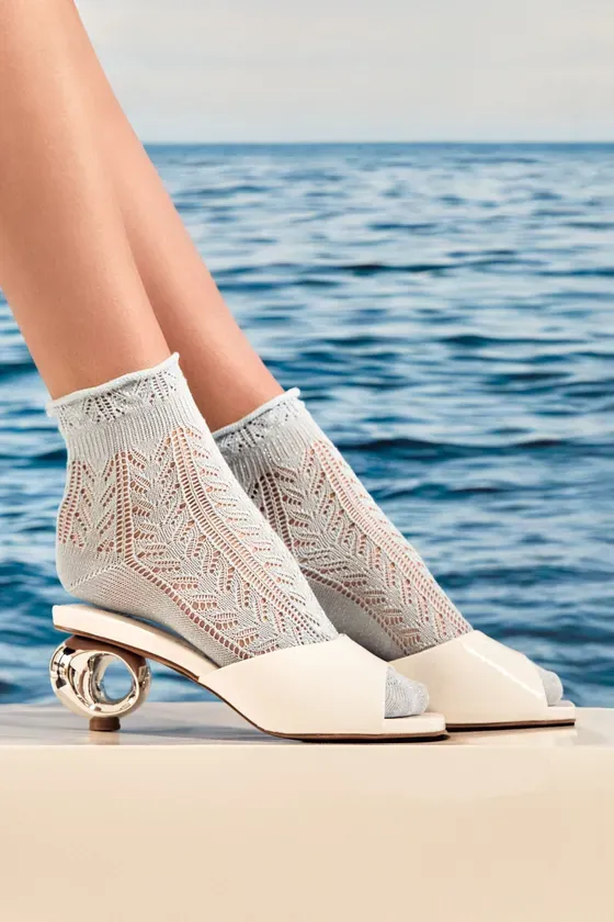 水辺で、白いサンダルと透かし模様の靴下を履いた女性の足のクローズアップ写真。背景には青い水面が見える。