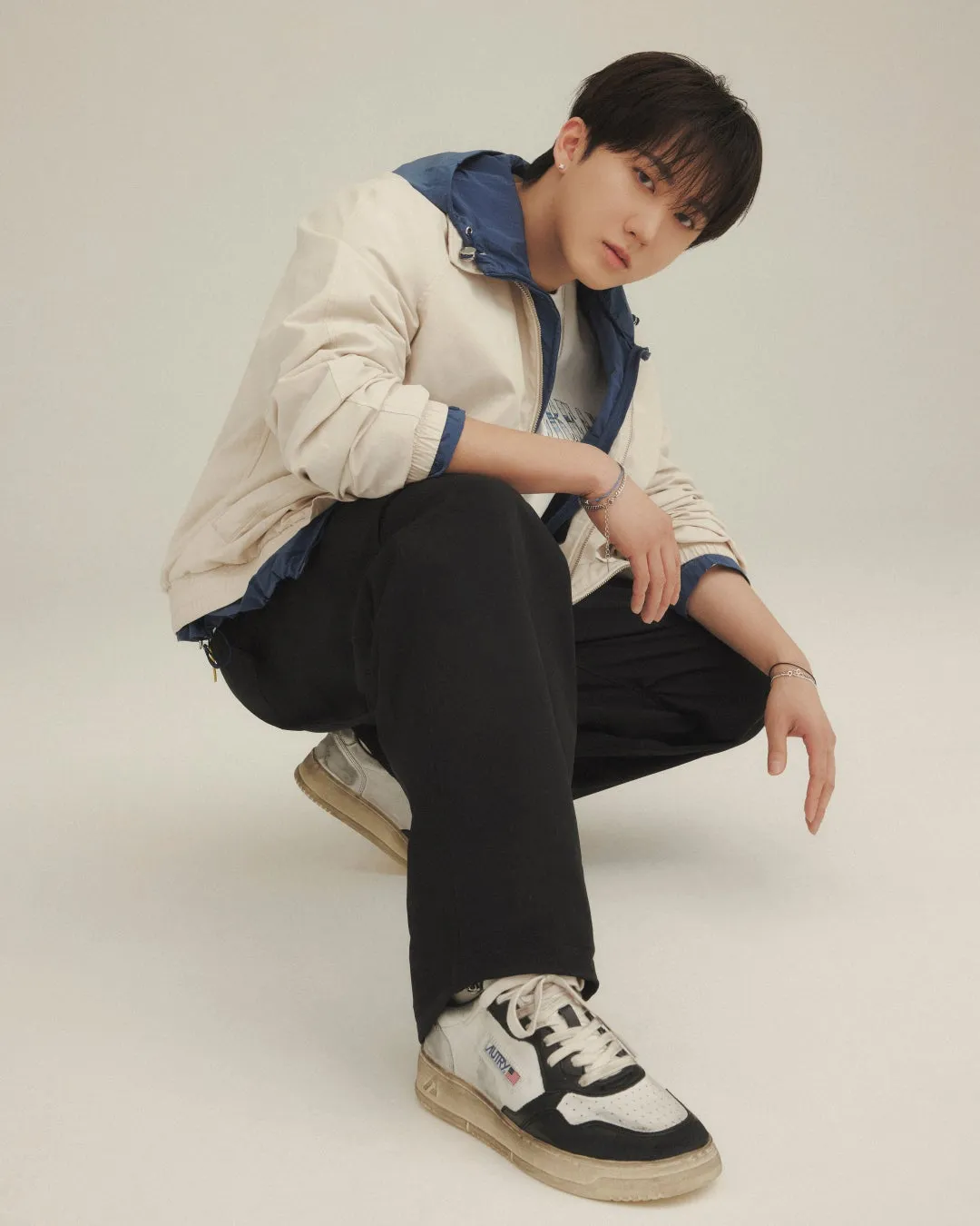 男性がしゃがみこんでカメラを見ている写真。彼は白いジャケットと黒いズボンを着ており、足元にはスニーカーを履いている。背景は白。