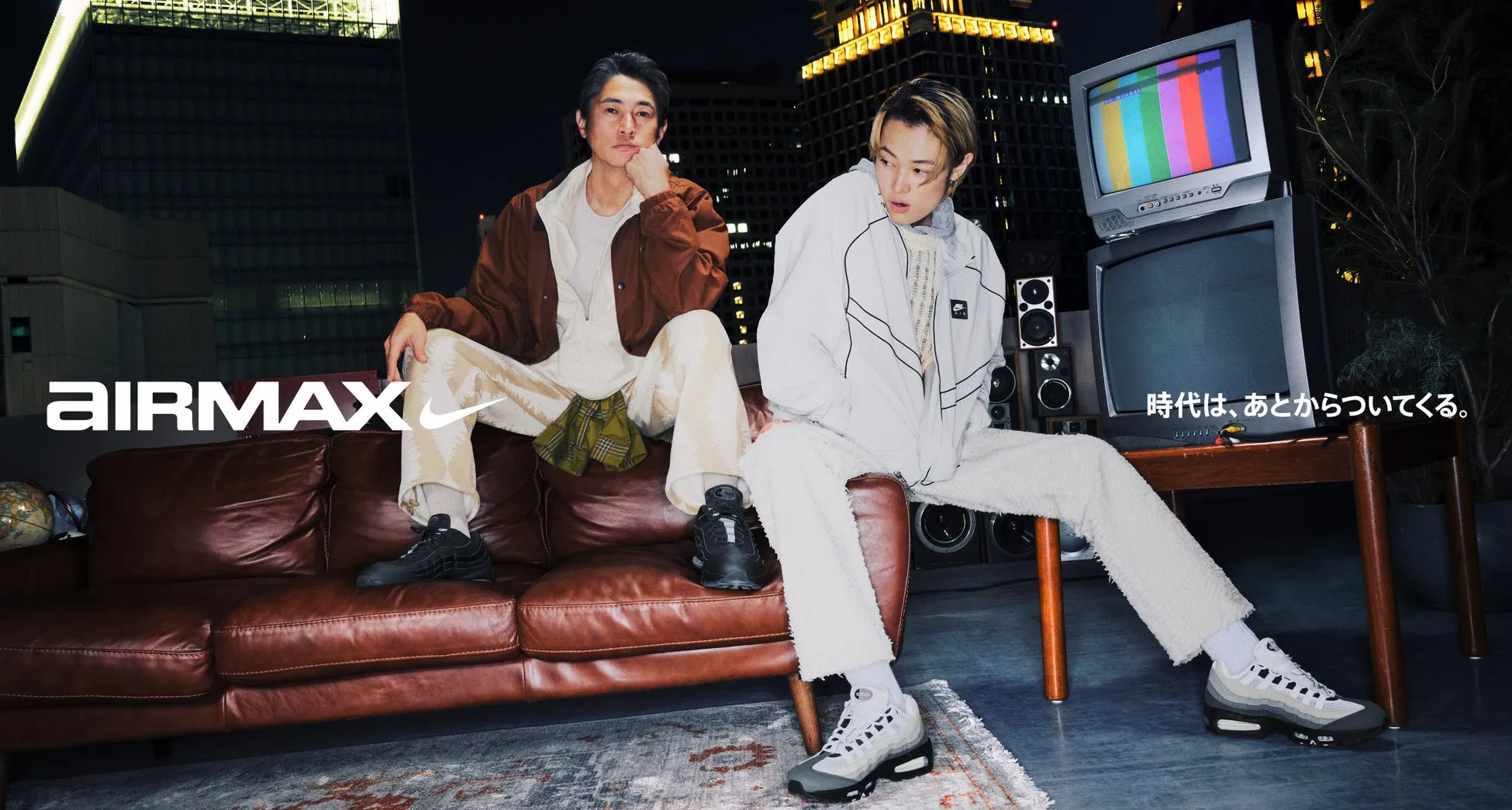 2人の男性がソファに座っており、ナイキのエアマックスの広告写真のようです。背景にはテレビ、スピーカー、都市の夜景が写っています。
